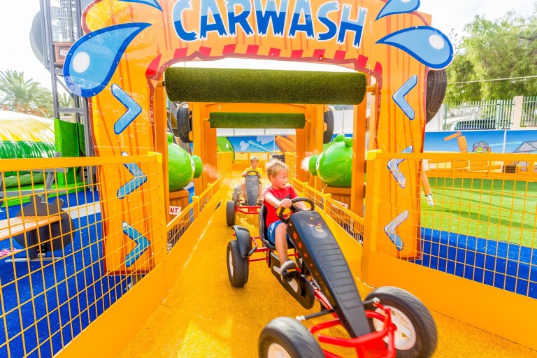 Child on a pedal cart under a colorful 'carwash' arch in a play area.