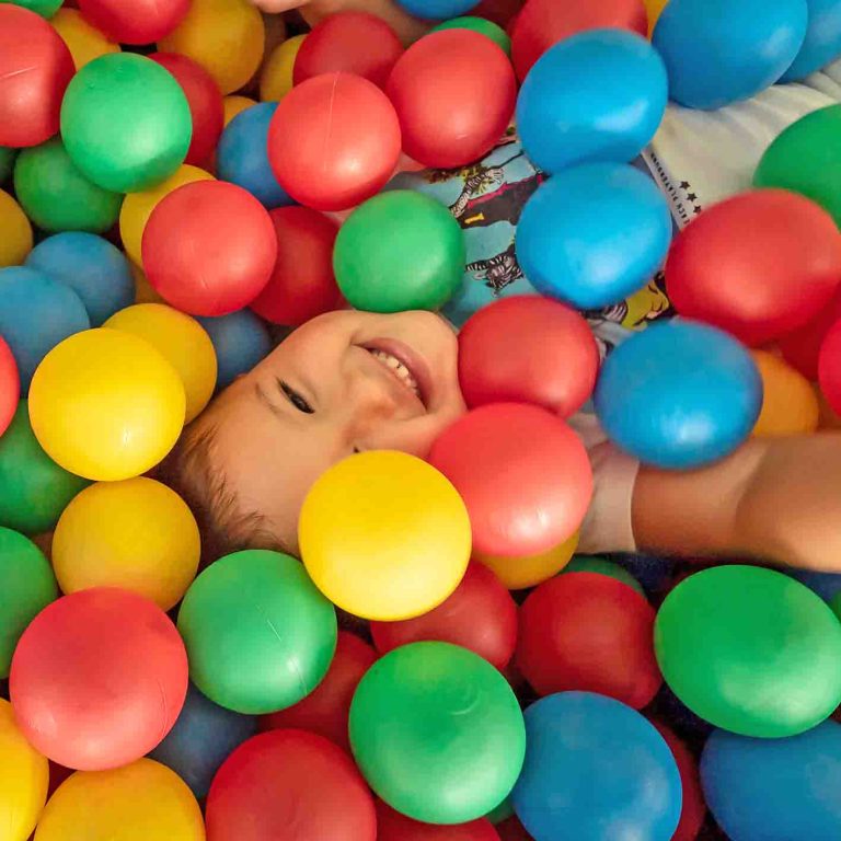Child smiling in a colorful ball pit, surrounded by red, blue, yellow, and green balls.