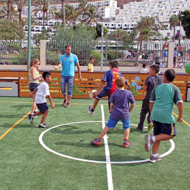 Children playing soccer outdoors on a small turf field with buildings in the background.