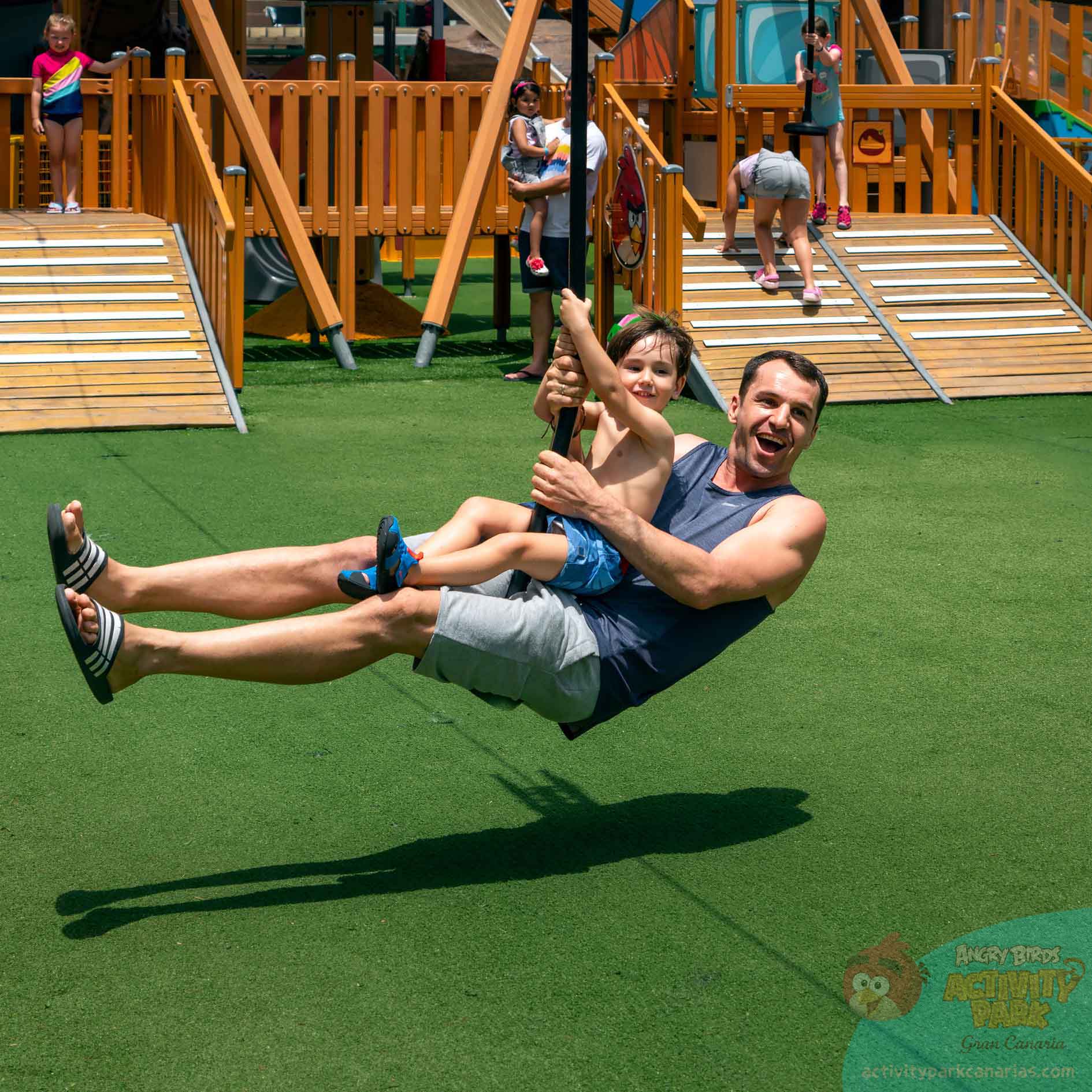 Man and child on zip line in playground with wooden structures.