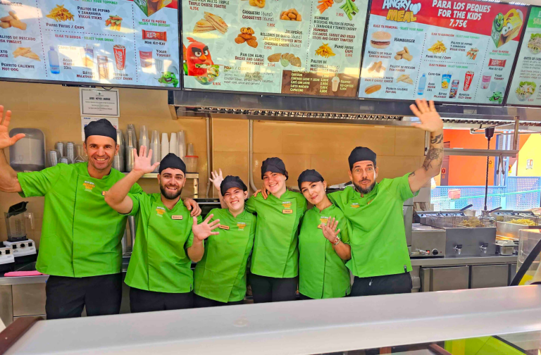 Group of smiling fast-food employees in green uniforms, waving beneath menu boards.