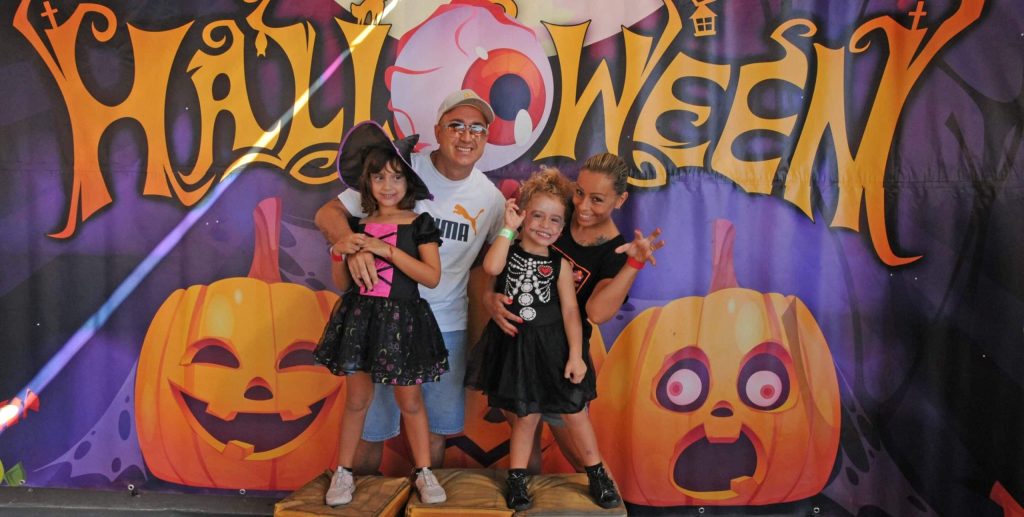 Group of four in costumes in front of Halloween backdrop with pumpkins and spooky text.