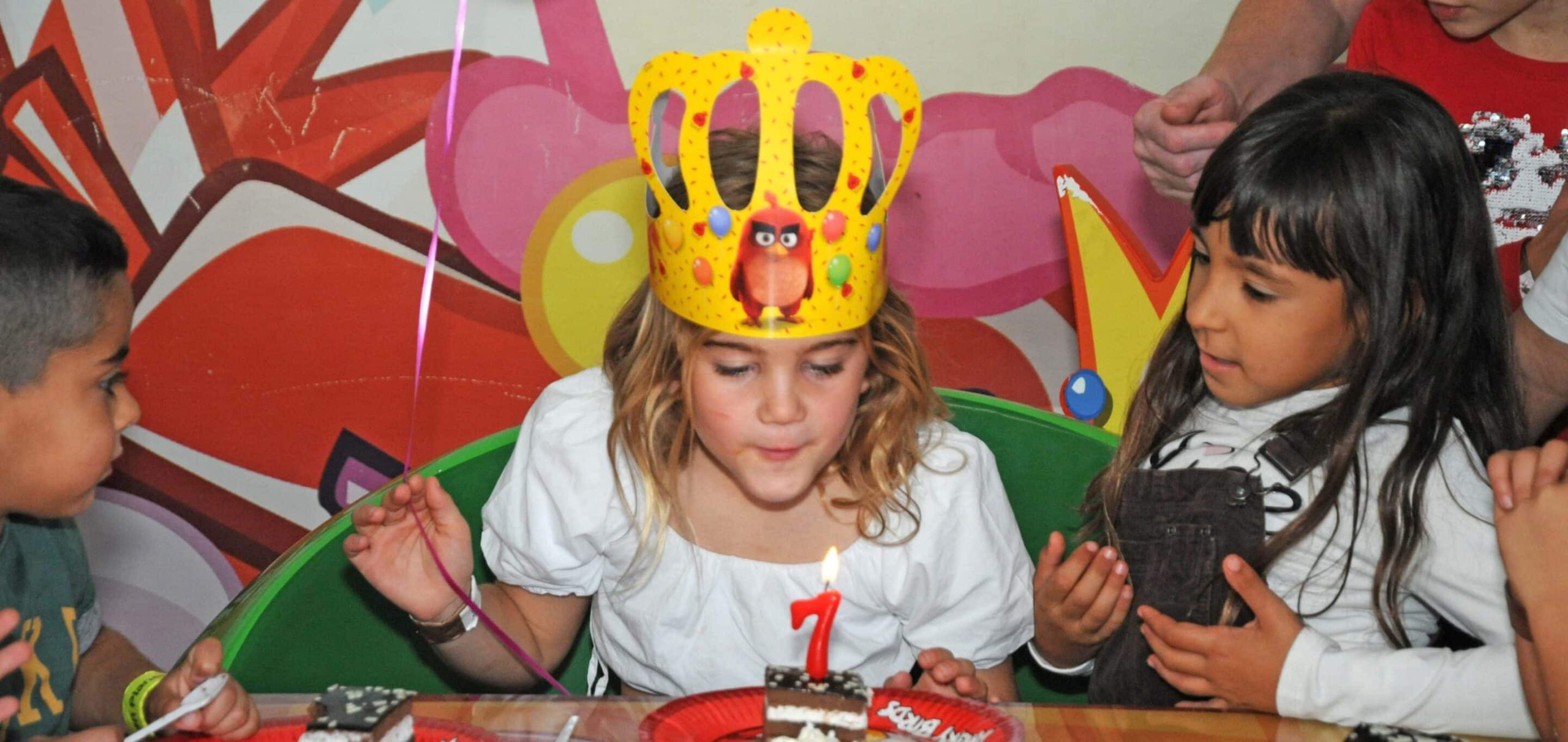 Child in crown blowing out candle on birthday cake with friends.
