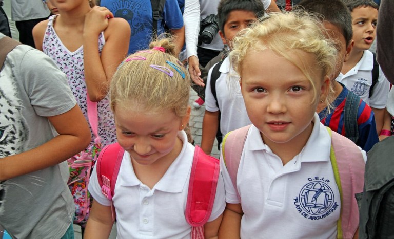 Two young children in school uniforms with backpacks in a crowd.